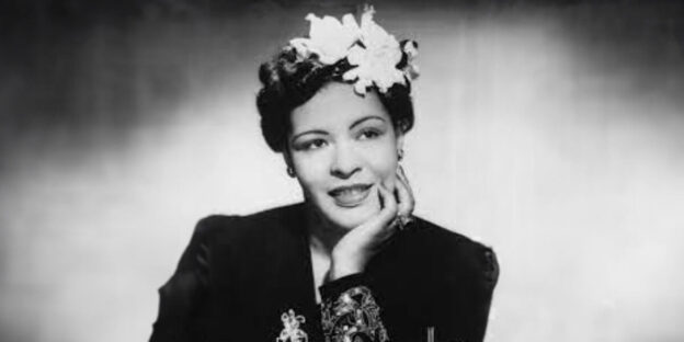 Black and white portrait of Billie Holiday resting her chin on her hand, smiling slightly, wearing a dark dress with floral embellishments and a large white flower in her hair.
