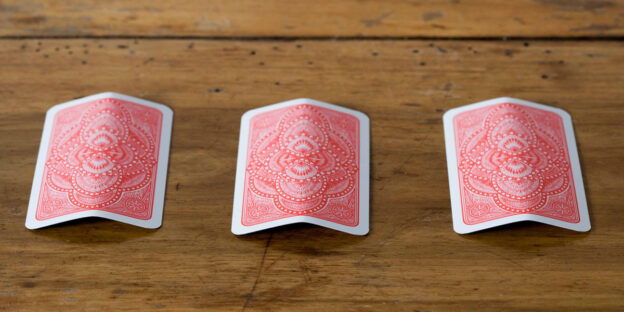 Three red-backed playing cards lie face-down on a wooden table, each slightly bent in the middle, arranged side-by-side as if set up for a classic shell game or a sleight-of-hand trick.