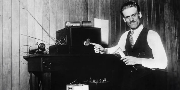 A black-and-white photo of an early 20th-century inventor in a vest and tie pointing at a cathode-ray tube on a bulky electronic device, surrounded by vintage equipment in a wood-paneled room.