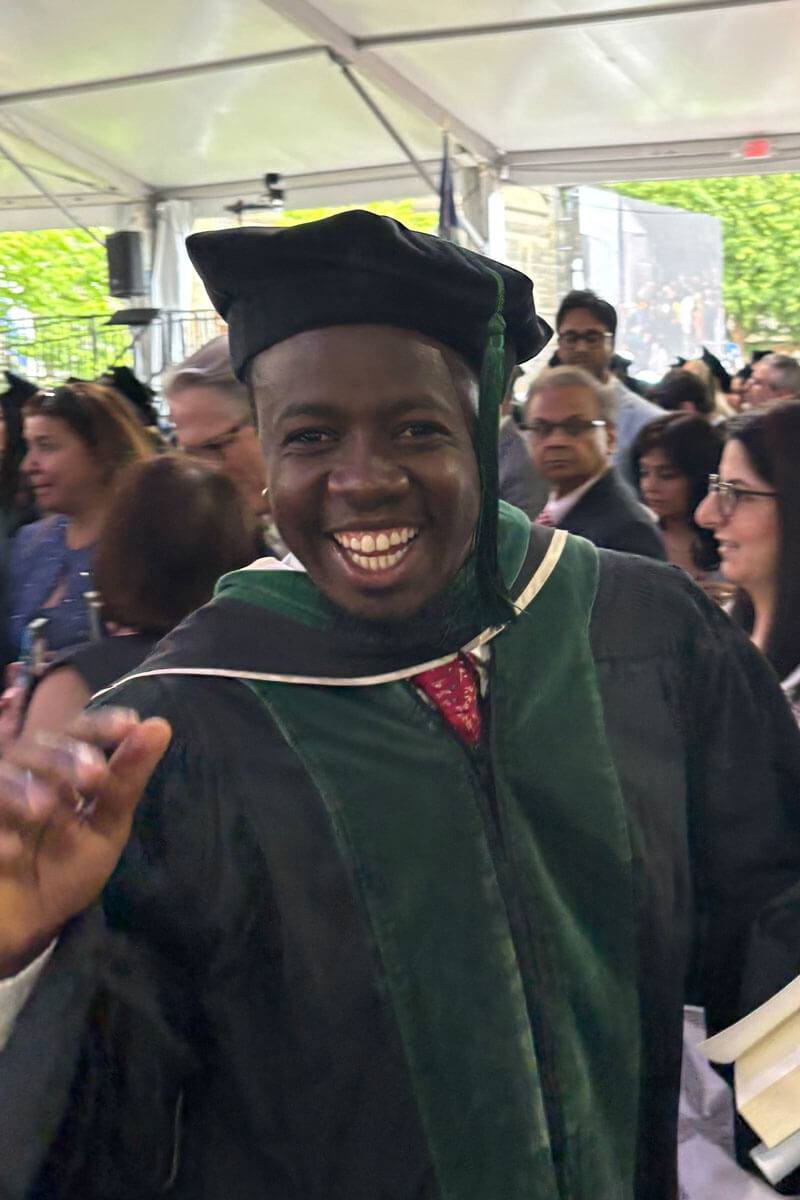 A smiling graduate in a green doctoral robe waves at the camera while surrounded by a crowd under a large graduation tent.