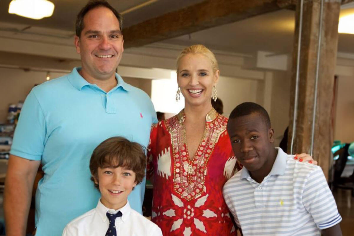 A smiling family stands indoors with two children, including a young boy in a white polo, gathered close and dressed for a celebration.