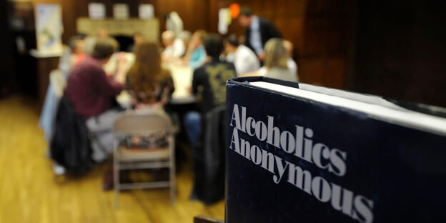 A close-up of a dark blue "Alcoholics Anonymous" book sits prominently in the foreground, with a blurred group of people gathered around a table in the background, suggesting a support meeting in a wood-paneled church basement.